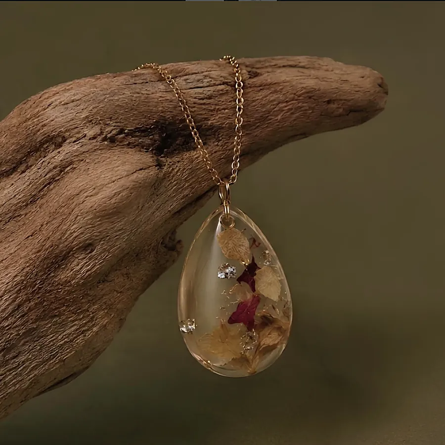 Ciondolo a goccia con petali chiari e cristalli, appoggiato su un legno levigato